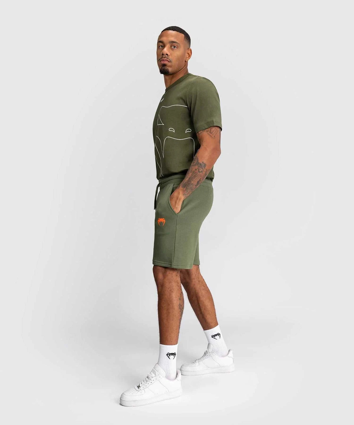 Man wearing khaki Shorts Venum Classic with elastic waistband, side and back pockets, and orange logo, paired with matching green t-shirt and white sneakers.