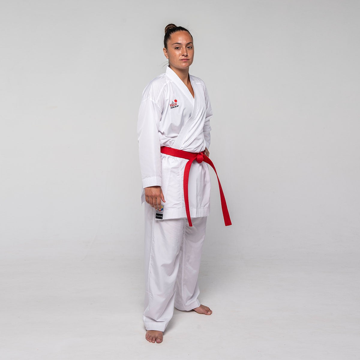 Professional karateka wearing white FUJIMAE Kumite ProWear Hyperlite QS WKF approved karate gi with red belt, standing against plain background
