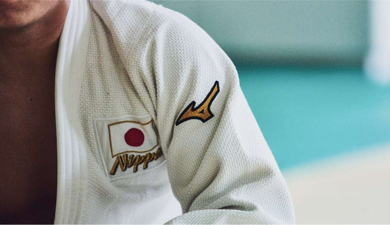 Gros plan d'un judoka portant un kimono blanc (judogi) avec le drapeau du Japon et le logo Mizuno sur l'épaule.