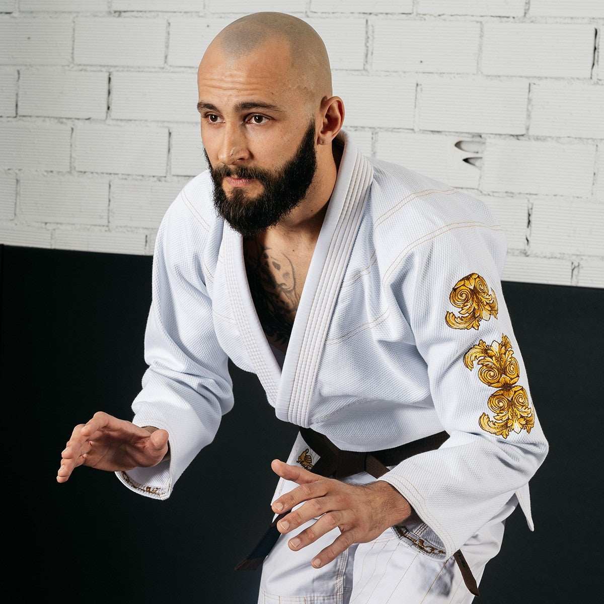 Man wearing white Shaka 21 BJJ Gi QS with gold embroidery on sleeve in a grappling stance against a white brick wall background
