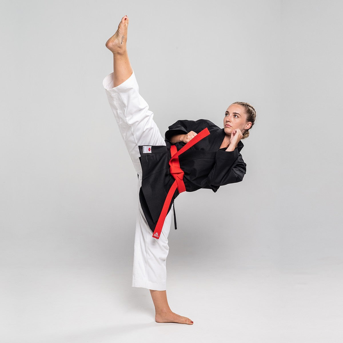 karate practitioner wearing white Pantalon Karaté Legacy II and black gi performing high kick against gray background