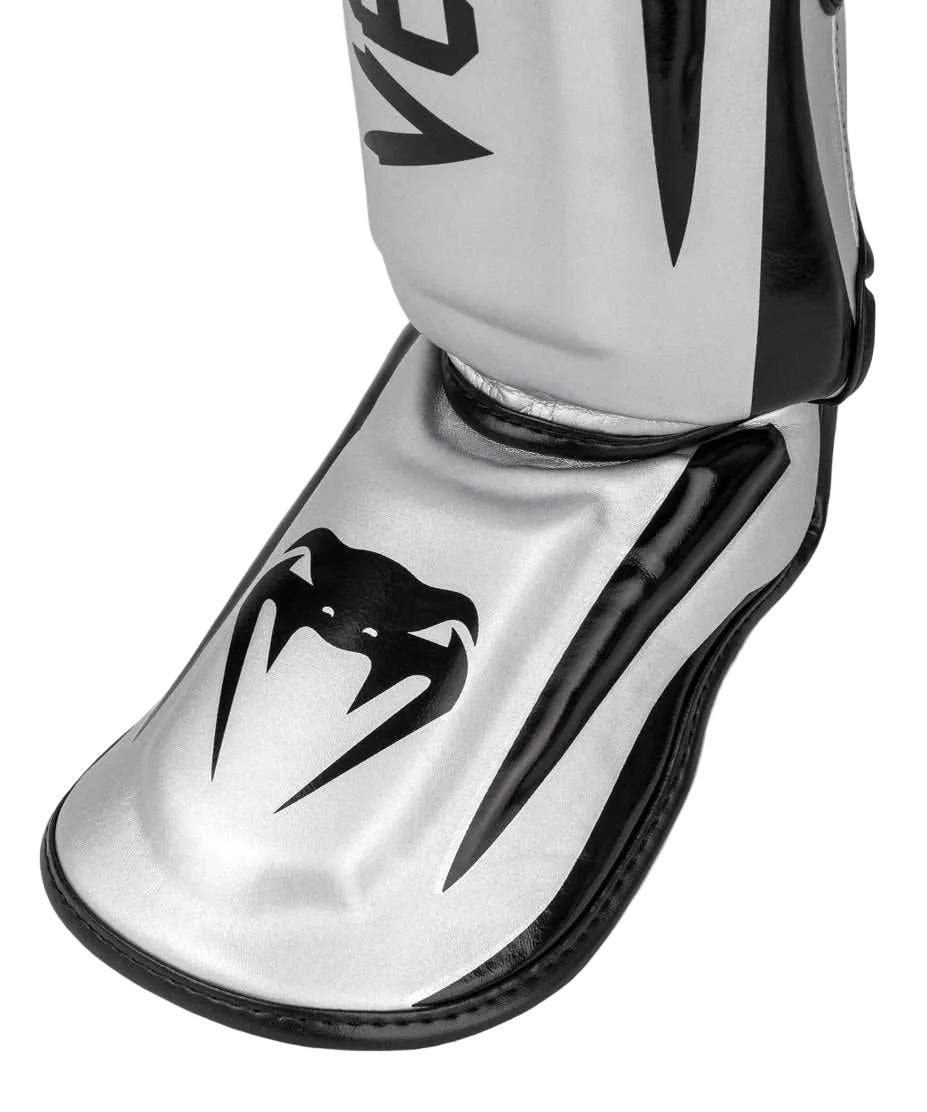 Protège-tibias Venum Elite argent noir en Skintex, absorption choc haute densité, maintien anatomique ajusté, fabrication thaïlandaise premium.