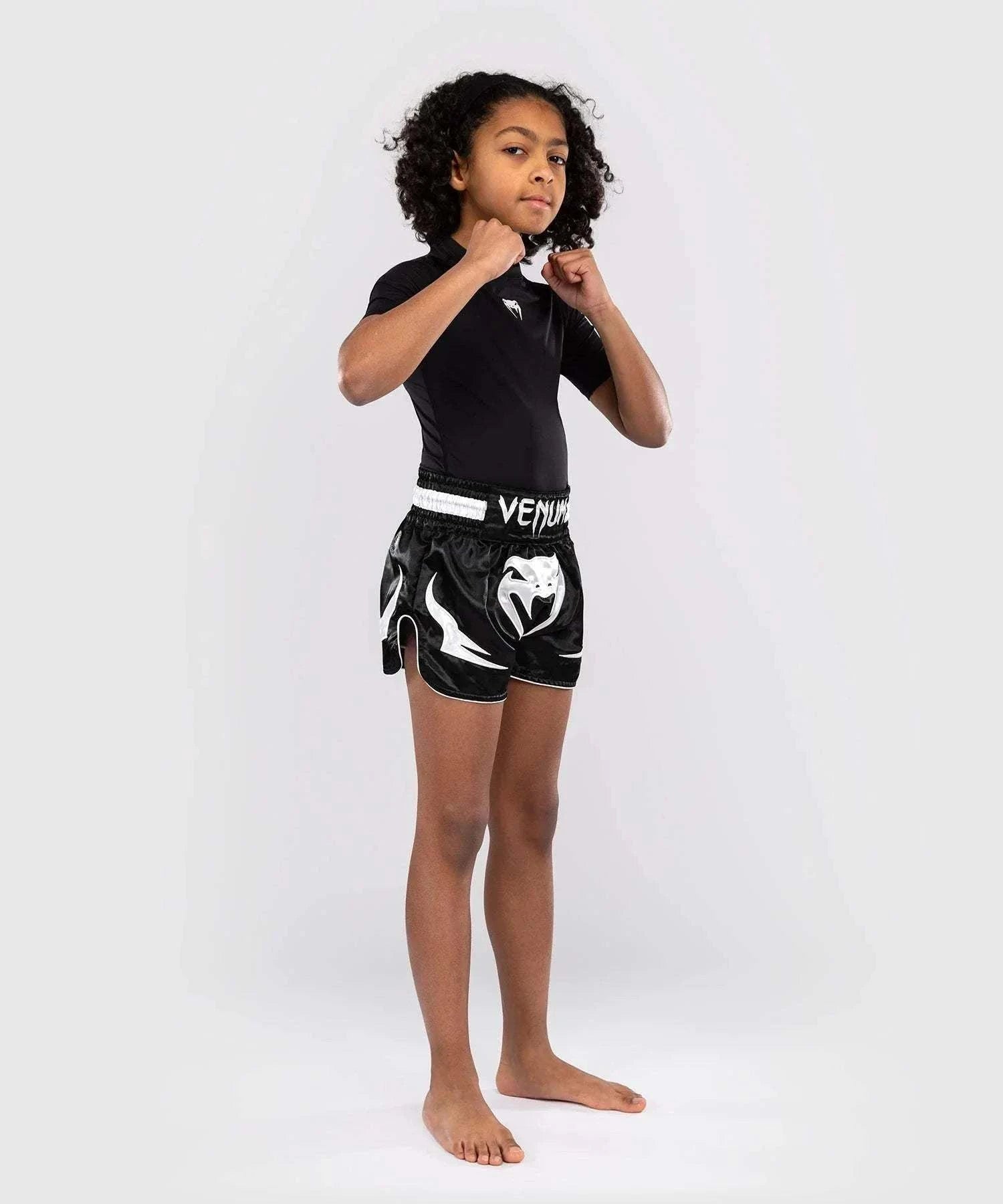 Child wearing black Short Muay Thai Venum Inferno with satin fabric and wide elastic waistband, in fighting stance