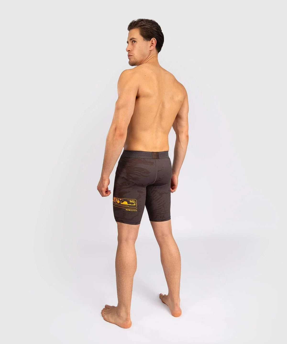 Male athlete wearing black UFC Fusion par Venum compression Vale Tudo fight shorts with yellow design on the side, standing barefoot facing away.