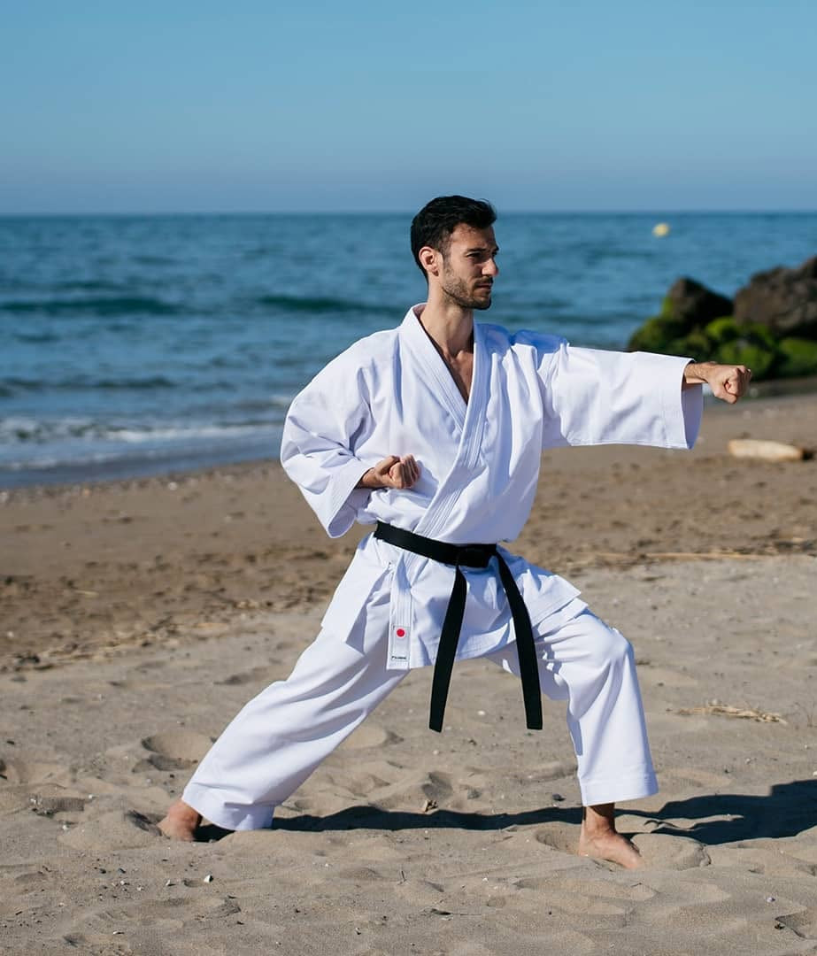 Un homme portant un kimono blanc (karatégi) et une ceinture noire effectue une posture de karaté (coup de poing vers l'avant) sur une plage de sable fin, face à la mer bleue en arrière-plan.