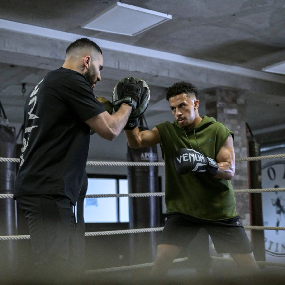 Un boxeur portant des gants noirs et une tenue verte s'entraîne sur un ring de boxe. Il lance un coup de poing sur la patte d'ours (cible de frappe) tenue par son entraîneur habillé en noir.