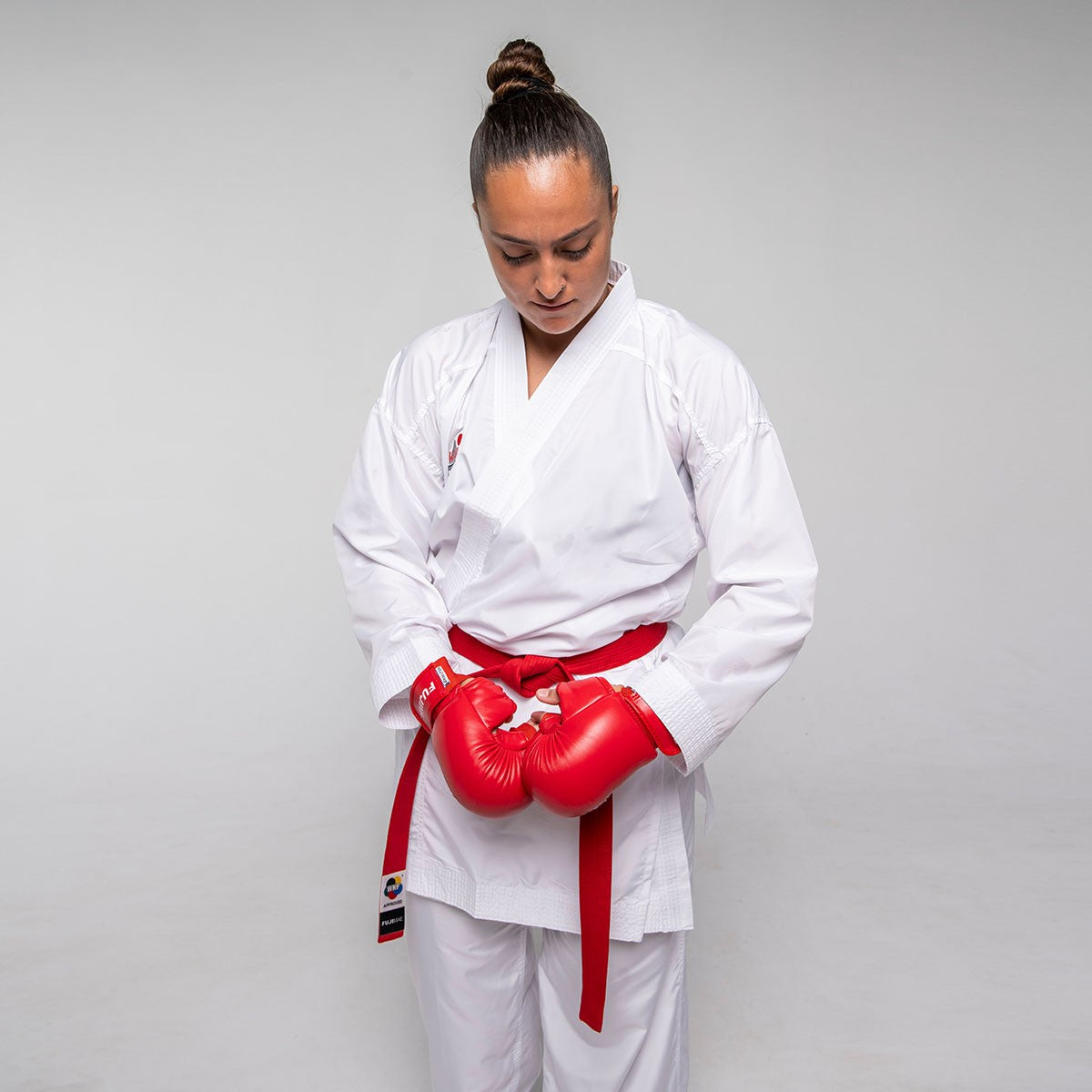 Female karate professional wearing white Kumite ProWear Hyperlite karate gi with red gloves adjusting belt against plain background